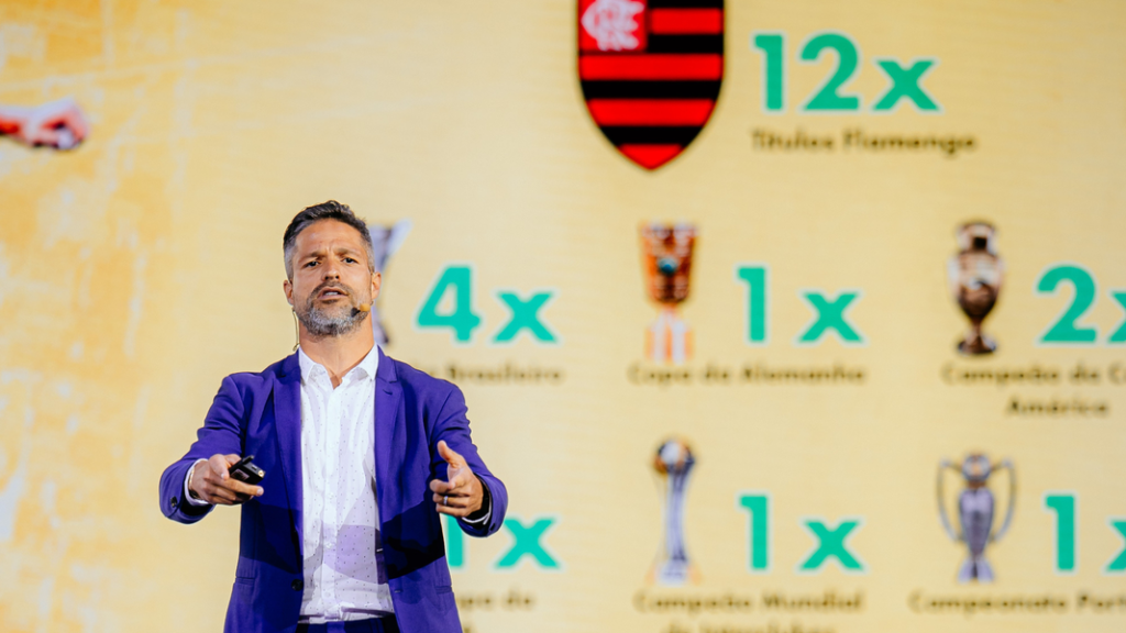 Homem vestindo um paletó azul sobre camisa branca, gesticula durante uma apresentação em um palco. Ao fundo, um telão amarelo exibe o escudo do Flamengo e uma lista de títulos conquistados, incluindo "12x Títulos Flamengo", "4x Brasileiro" e "1x Copa da Alemanha", acompanhados por ícones de troféus.