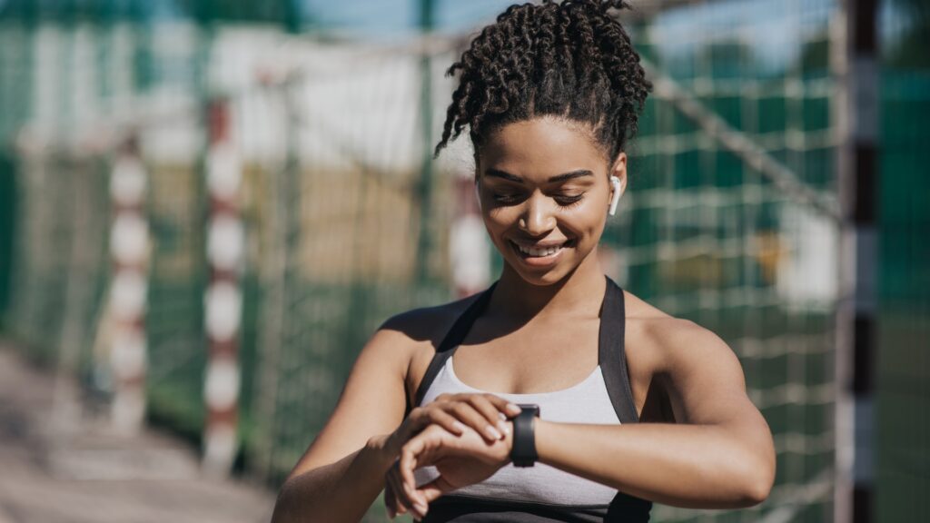 Mulher jovem, com cabelos presos em um coque alto, sorri enquanto olha para um smartwatch em seu pulso. Ela veste roupa esportiva branca e preta e usa fones de ouvido sem fio. O cenário ao fundo é um campo esportivo iluminado pelo sol, com redes de proteção desfocadas.