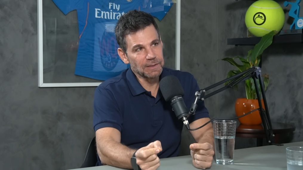 Homem de meia-idade, com cabelos grisalhos e barba curta, veste uma camisa polo azul-marinho enquanto fala em um microfone profissional de podcast. Ele está sentado em uma mesa com um copo de água à frente. Ao fundo, a parede cinza exibe uma camisa de futebol azul emoldurada e uma prateleira com uma bola de tênis amarela gigante e uma planta.